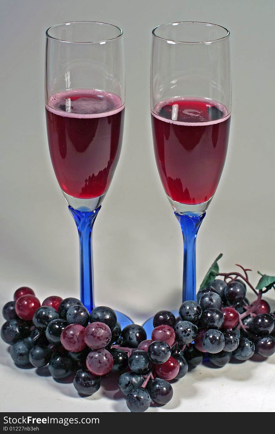 Two glasses of red wine in blue-stemmed glasses isolated. Red grapes cover the bottoms of the glasses. Two glasses of red wine in blue-stemmed glasses isolated. Red grapes cover the bottoms of the glasses.