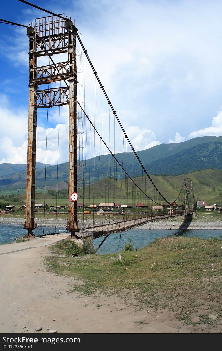 Hinged bridge through the river, on a background of mountains under clouds