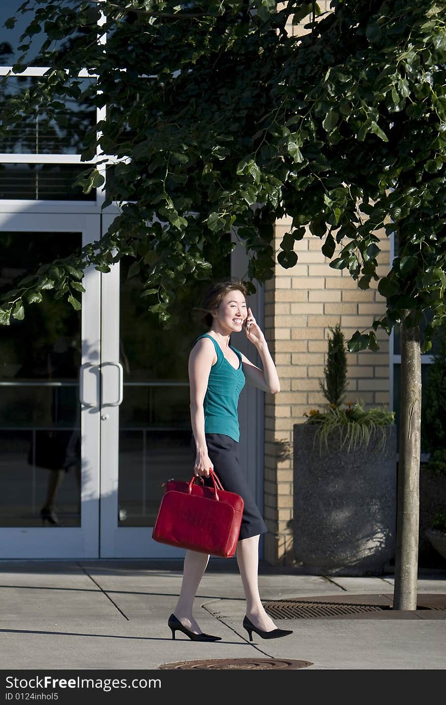 Young woman runs along building while talking on a cell phone. She is wearing a tank top and carrying a red bag. Vertically framed photo. Young woman runs along building while talking on a cell phone. She is wearing a tank top and carrying a red bag. Vertically framed photo.