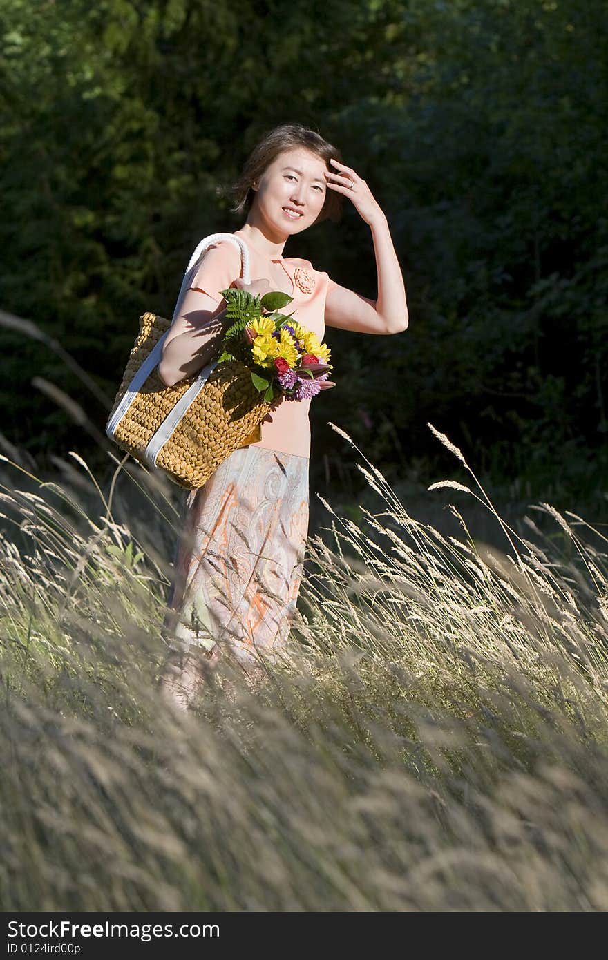 Attractive woman stands in a field while holding a basket containing a bouquet of flowers. She is standing to the side, smiling and brushing hair from her face.  Vertically framed photo. Attractive woman stands in a field while holding a basket containing a bouquet of flowers. She is standing to the side, smiling and brushing hair from her face.  Vertically framed photo.