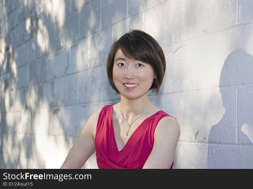 Young Asian woman sits against white brick wall. Woman is smiling at camera. Horizontally framed photo. Young Asian woman sits against white brick wall. Woman is smiling at camera. Horizontally framed photo.