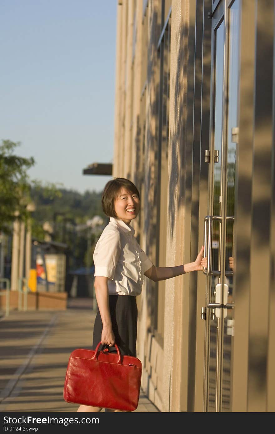 Young woman smiles at camera while opening a door to a building. She is smiling at the camera and carrying a red bag. Vertically framed photo. Young woman smiles at camera while opening a door to a building. She is smiling at the camera and carrying a red bag. Vertically framed photo.