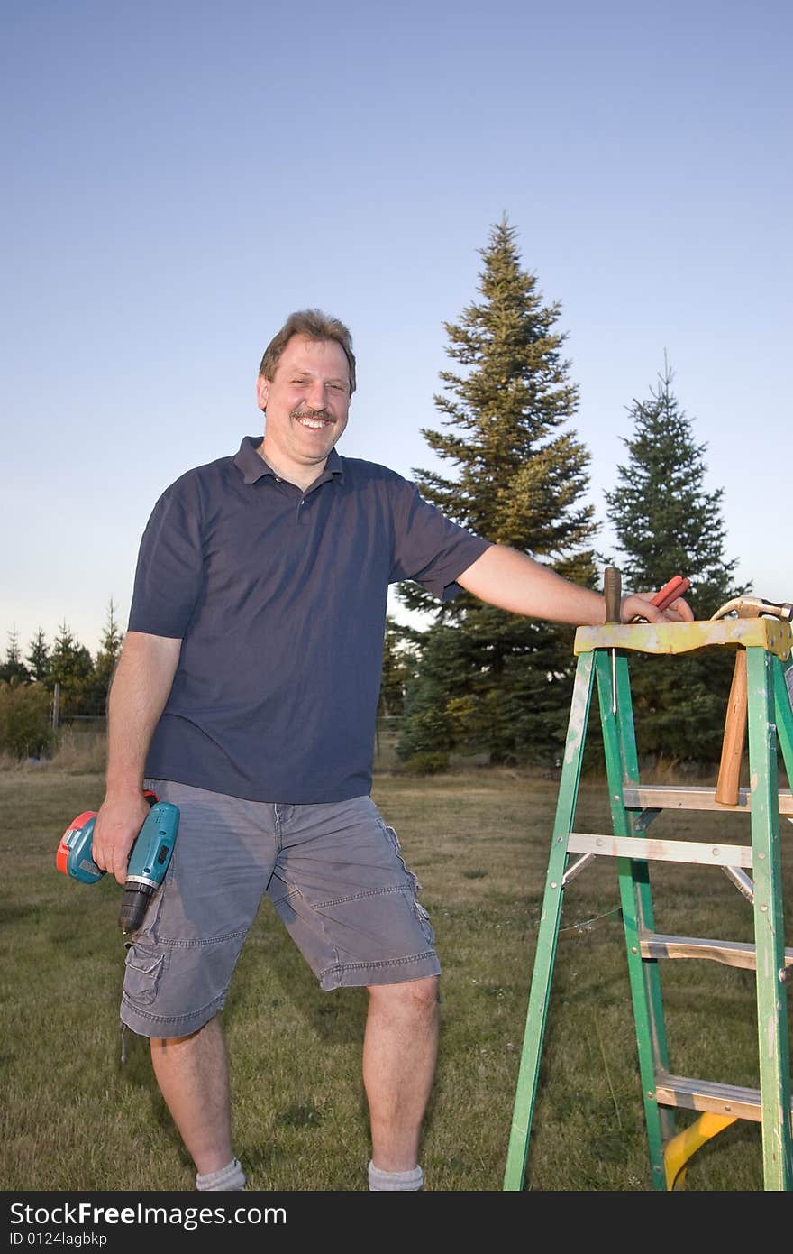 Man standing next to a ladder holding a drill and smiling. Vertically framed photograph. Man standing next to a ladder holding a drill and smiling. Vertically framed photograph