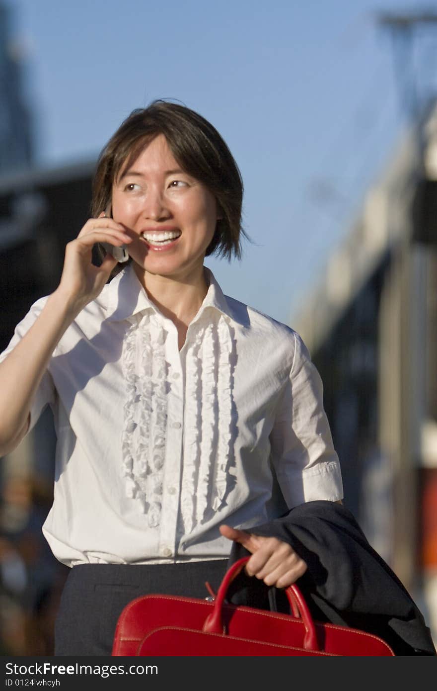 Young woman walks while talking on her cell phone. She is smiling and carrying a red bag. Vertically framed photo. Young woman walks while talking on her cell phone. She is smiling and carrying a red bag. Vertically framed photo.