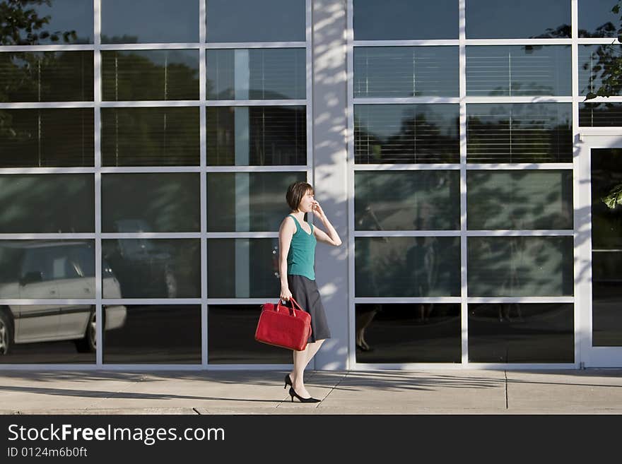 Young woman walks along building while talking on a cell phone and carrying a red bag. She is wearing a tank top. Horizontally framed photo. Young woman walks along building while talking on a cell phone and carrying a red bag. She is wearing a tank top. Horizontally framed photo.