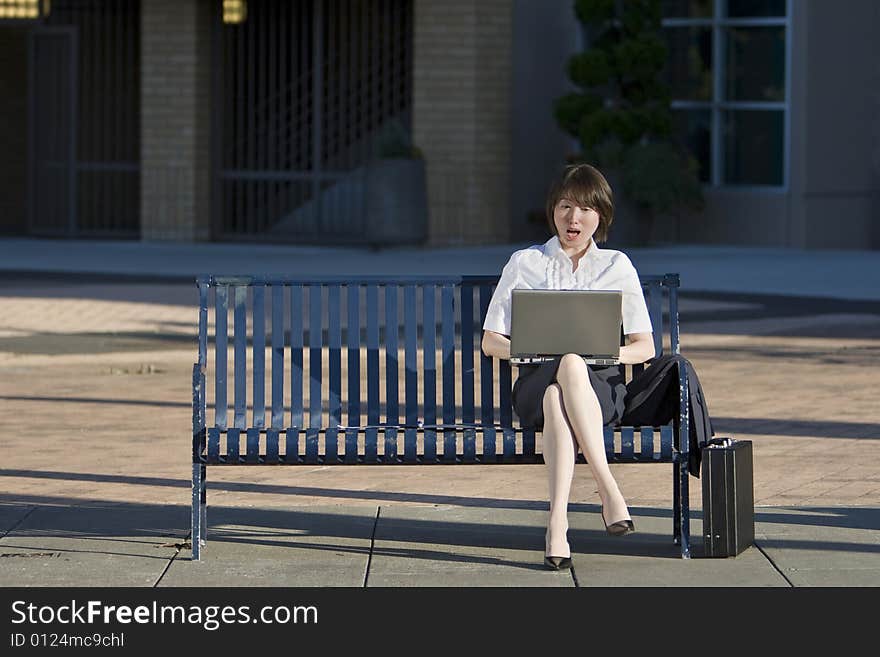 Young woman sits on a bench in front of a building. She is looking at her laptop and looks surprised. Vertically framed photo. Young woman sits on a bench in front of a building. She is looking at her laptop and looks surprised. Vertically framed photo.