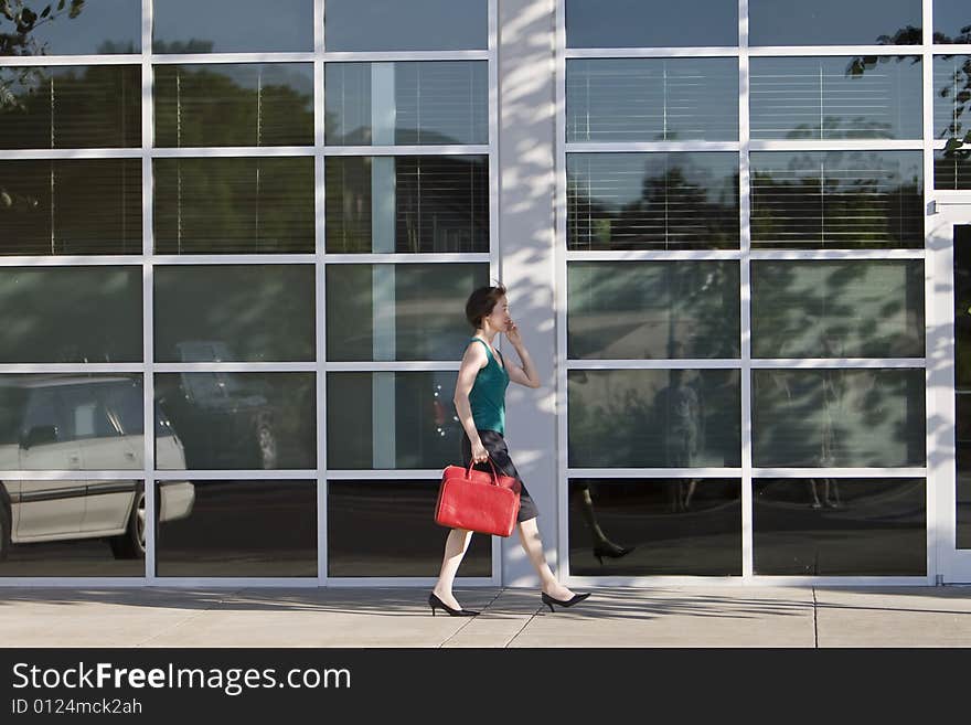 Young woman walks along building while talking on a cell phone and carrying a red bag. She is wearing a tank top. Horizontally framed photo. Young woman walks along building while talking on a cell phone and carrying a red bag. She is wearing a tank top. Horizontally framed photo.