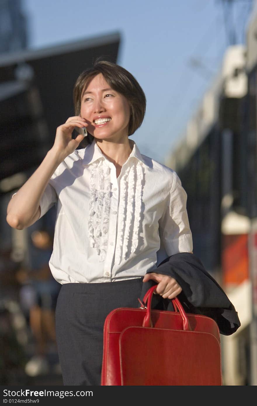 Young woman walks while talking on her cell phone. She is carrying a red bag. Vertically framed photo. Young woman walks while talking on her cell phone. She is carrying a red bag. Vertically framed photo.