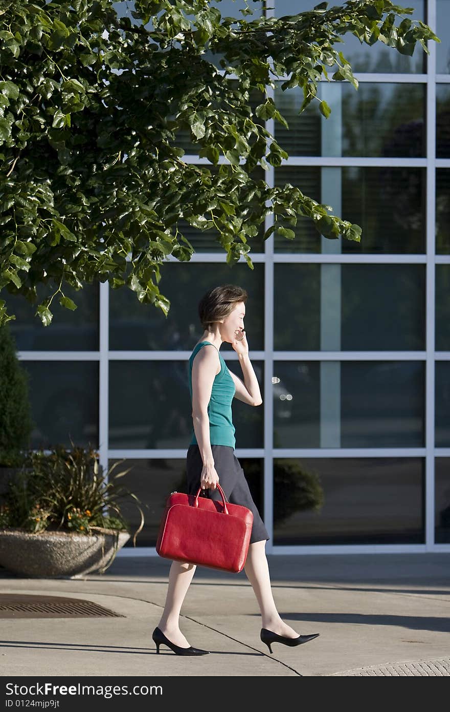 Young woman runs along building while talking on a cell phone. She is wearing a tank top and carrying a red bag. Vertically framed photo. Young woman runs along building while talking on a cell phone. She is wearing a tank top and carrying a red bag. Vertically framed photo.