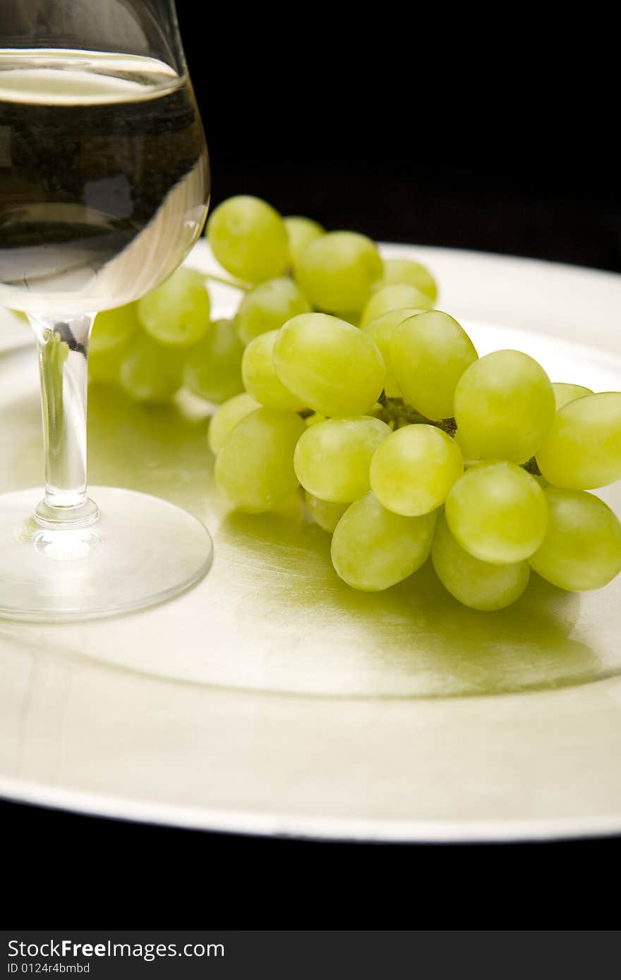 White wine and white grapes on a silver tray. Black background. Focus on grapes. White wine and white grapes on a silver tray. Black background. Focus on grapes