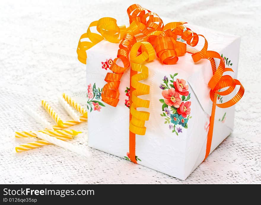 Beautiful gift box with vibrant orange and yellow ribbons sitting on a white lace tablecloth with birthday candles. Beautiful gift box with vibrant orange and yellow ribbons sitting on a white lace tablecloth with birthday candles.