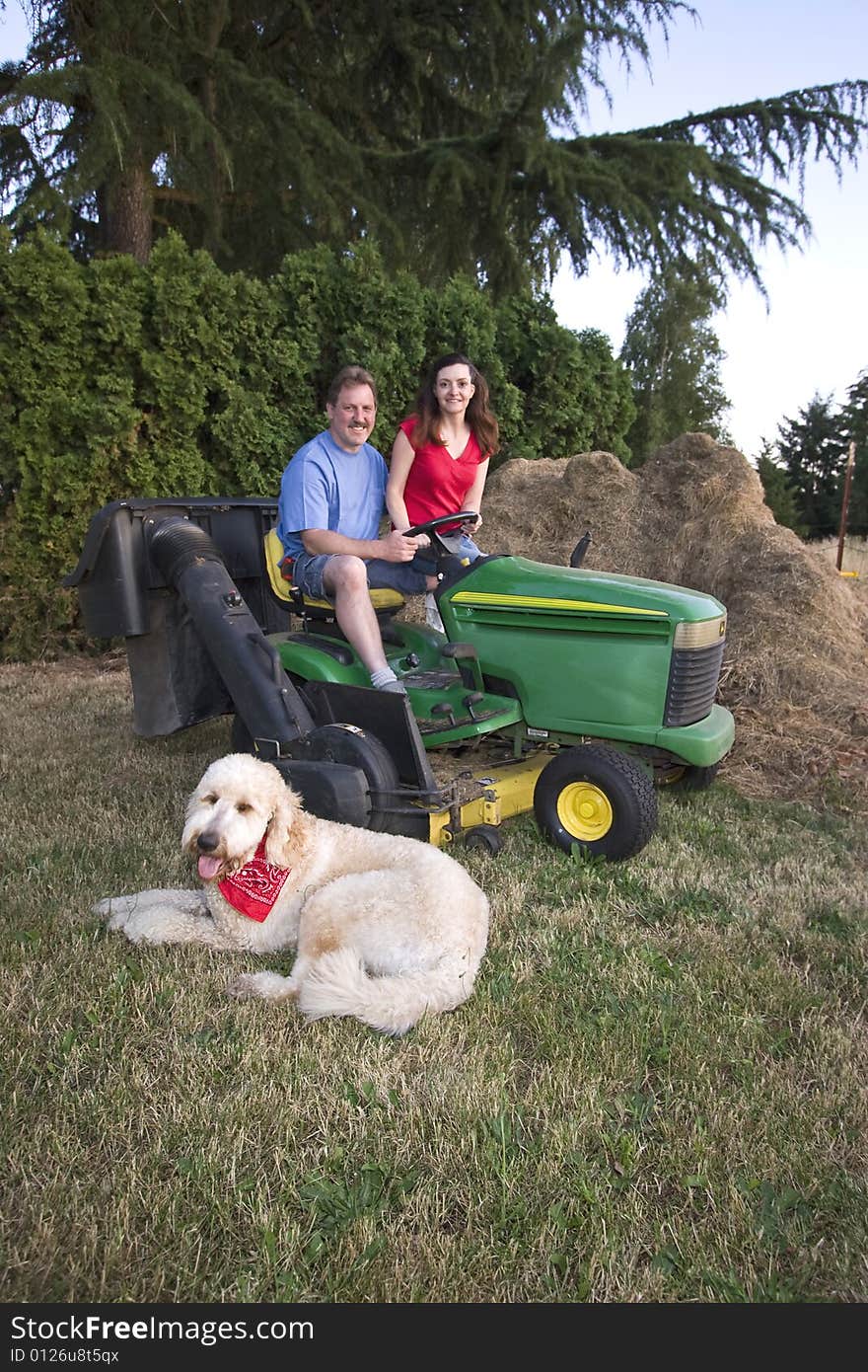 A man and woman sitting on a tractor smiling. Their dog is nearby on the grass. Vertically framed photograph. A man and woman sitting on a tractor smiling. Their dog is nearby on the grass. Vertically framed photograph