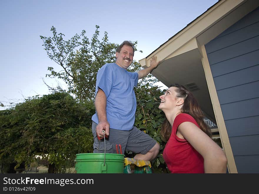 Man smiling as he stands on a ladder holding a bucket. His wife smiles up at him. Horizontally framed photograph. Man smiling as he stands on a ladder holding a bucket. His wife smiles up at him. Horizontally framed photograph