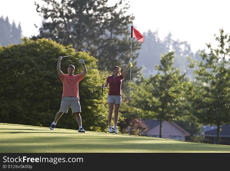 A young couple is playing on a golf course.  The man is holding his golf club above his head and the woman is holding the flag.  They are smiling at the camera.  Horizontally framed shot. A young couple is playing on a golf course.  The man is holding his golf club above his head and the woman is holding the flag.  They are smiling at the camera.  Horizontally framed shot.