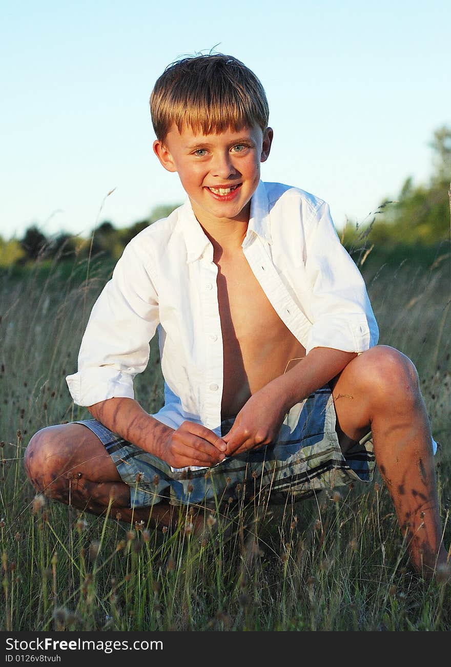 A young boy is sitting outside in a field of grass.  He is smiling at the camera.  Vertically framed shot. A young boy is sitting outside in a field of grass.  He is smiling at the camera.  Vertically framed shot.