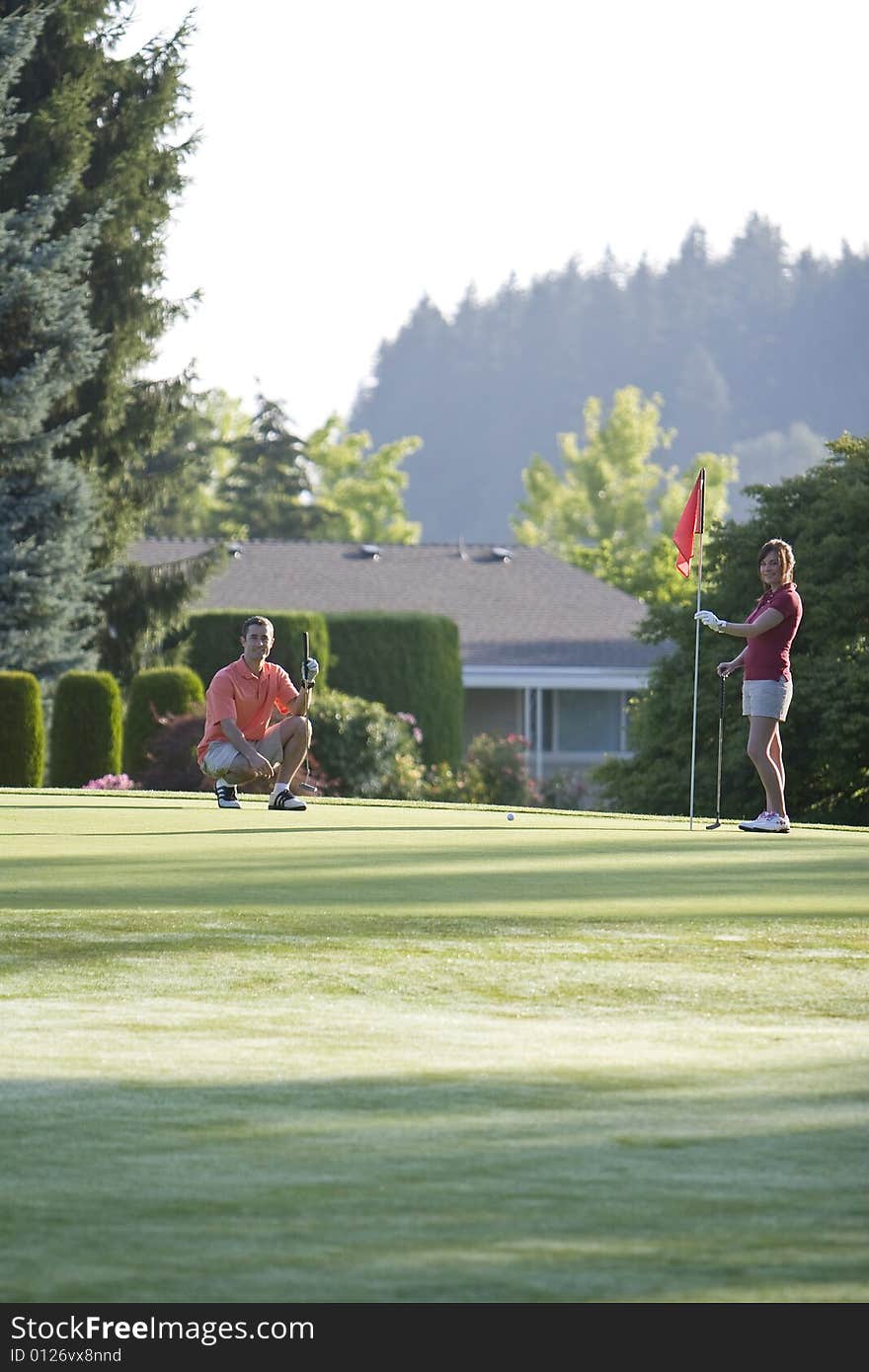 A young couple is setting up to play golf on the green of a golf course.  They are looking at the camera.  Vertically framed shot. A young couple is setting up to play golf on the green of a golf course.  They are looking at the camera.  Vertically framed shot.