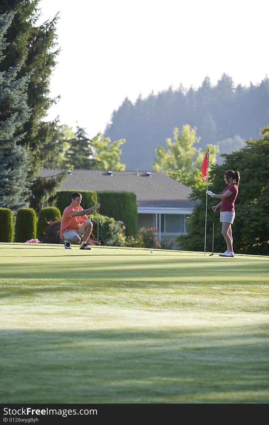 A young couple is setting up to play golf on the green of a golf course.  They are looking away from the camera.  Vertically framed shot. A young couple is setting up to play golf on the green of a golf course.  They are looking away from the camera.  Vertically framed shot.