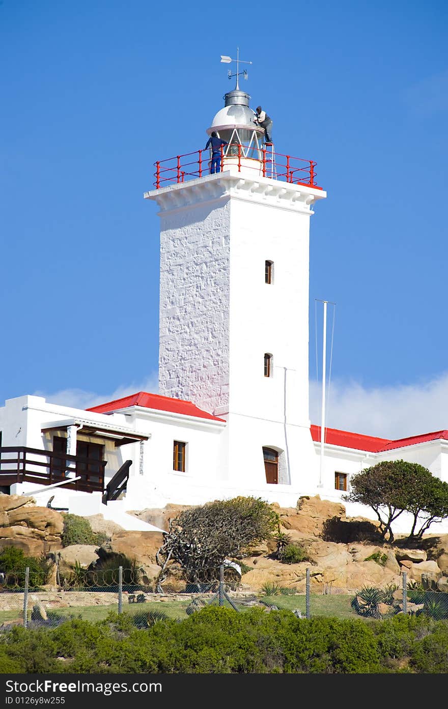 A newly renovated lighthouse and weather station against a bright blue sky. Two workers are visible, busy painting the dome.