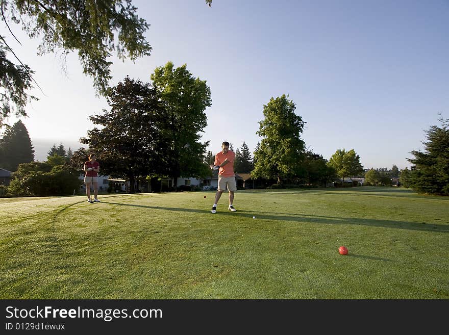 A young couple is standing on a golf course and playing golf.  The man is looking down at the ball he is swinging at and the woman is looking at the man.  Horizontally framed shot. A young couple is standing on a golf course and playing golf.  The man is looking down at the ball he is swinging at and the woman is looking at the man.  Horizontally framed shot.