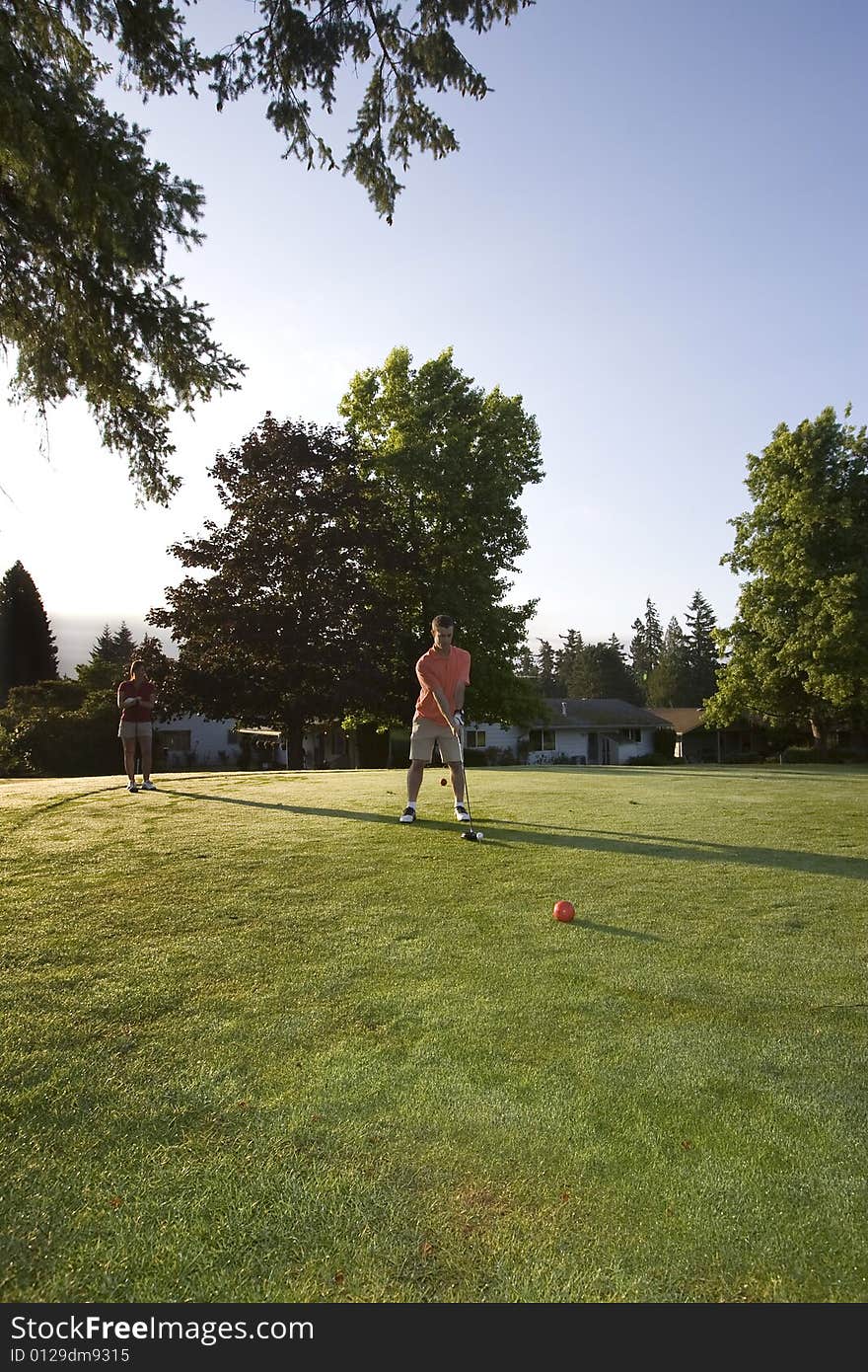 A young couple is standing on a golf course and playing golf.  The man is looking down at the ball he is swinging at and the woman is looking at the man.  Vertically framed shot. A young couple is standing on a golf course and playing golf.  The man is looking down at the ball he is swinging at and the woman is looking at the man.  Vertically framed shot.