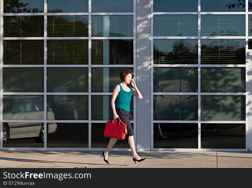 Yound Asian woman walks briskly while talking on the cell phone.  She is carrying a red handbag. Horizontally framed photo. Yound Asian woman walks briskly while talking on the cell phone.  She is carrying a red handbag. Horizontally framed photo.