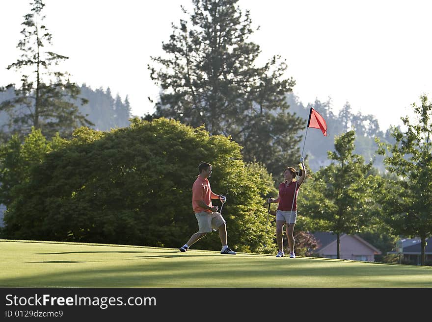 A young couple is playing on a golf course.  The man is holding his golf club and the woman is holding the flag.  They are smiling and looking away from the camera.  Horizontally framed shot. A young couple is playing on a golf course.  The man is holding his golf club and the woman is holding the flag.  They are smiling and looking away from the camera.  Horizontally framed shot.