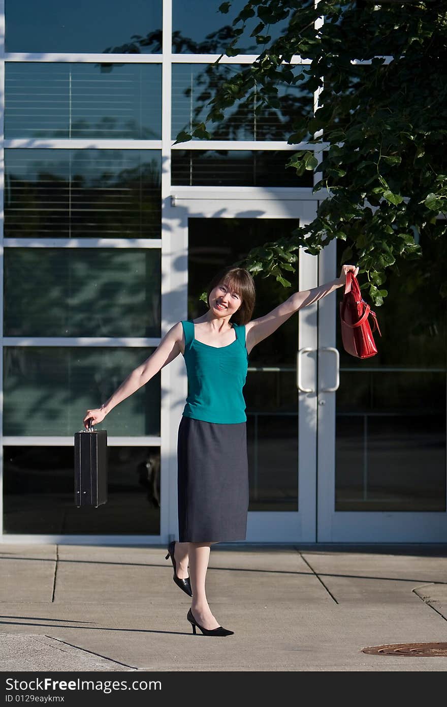 Woman smiles at camera while holding briefcase and red handbag out to her side. She is kicking up one leg. Vertically framed photo. Woman smiles at camera while holding briefcase and red handbag out to her side. She is kicking up one leg. Vertically framed photo.