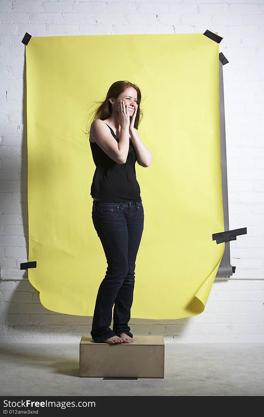 Young red headed woman standing on wooden box in front of yellow paper background laughs so much her cheeks hurt. Young red headed woman standing on wooden box in front of yellow paper background laughs so much her cheeks hurt.