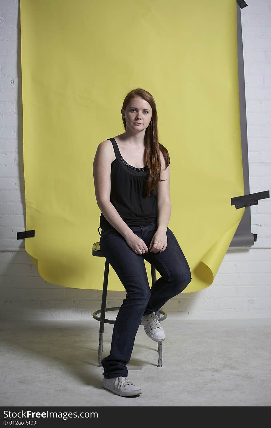 Young red headed woman sitting on a metal stool in front of a yellow background. Young red headed woman sitting on a metal stool in front of a yellow background.