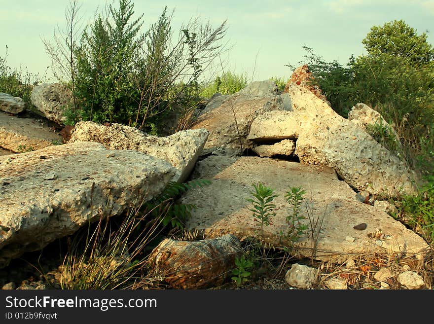 A pile of large boulders and small rocks in a field, with grass and weeds growing between them, against a cloudy blue sky. A pile of large boulders and small rocks in a field, with grass and weeds growing between them, against a cloudy blue sky