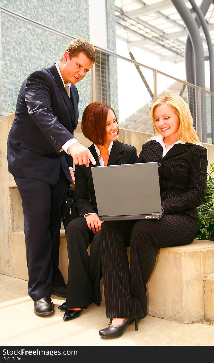 Two business women sitting outdoor holding laptop and another business man pointing at the laptop. Two business women sitting outdoor holding laptop and another business man pointing at the laptop