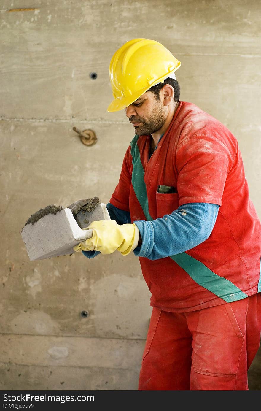 Construction worker holds cinder block while layering cement. He is wearing an orange suit and a yellow hard hat. Vertically framed photo. Construction worker holds cinder block while layering cement. He is wearing an orange suit and a yellow hard hat. Vertically framed photo.