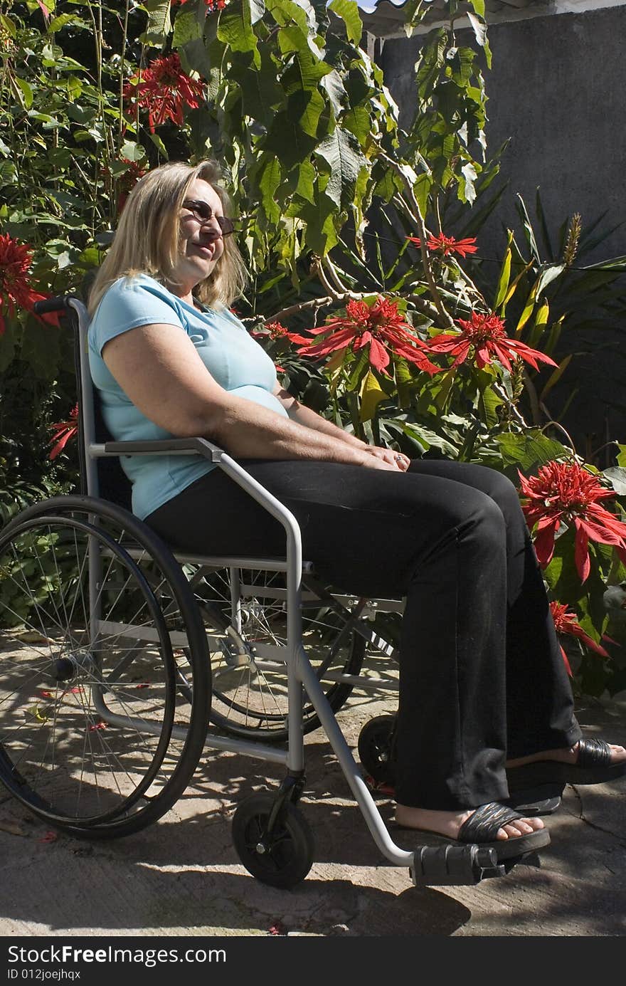 Woman in blue shirt reclines in wheelchair next to flowery bush. She is wearing sunglasses and looking ahead. Vertically framed photo. Woman in blue shirt reclines in wheelchair next to flowery bush. She is wearing sunglasses and looking ahead. Vertically framed photo.