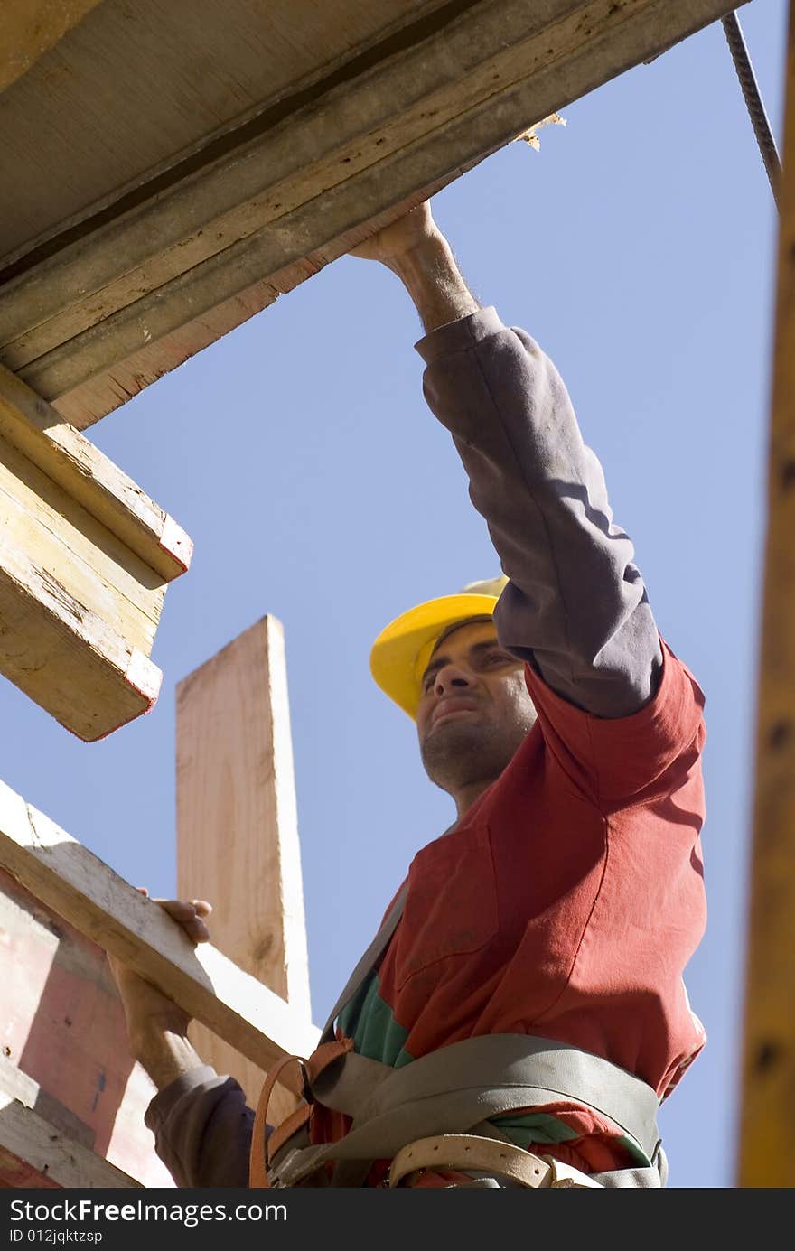 Construction worker carries board up ladder. He is wearing an orange suit and a yellow hard hat. Vertically framed photo. Construction worker carries board up ladder. He is wearing an orange suit and a yellow hard hat. Vertically framed photo.