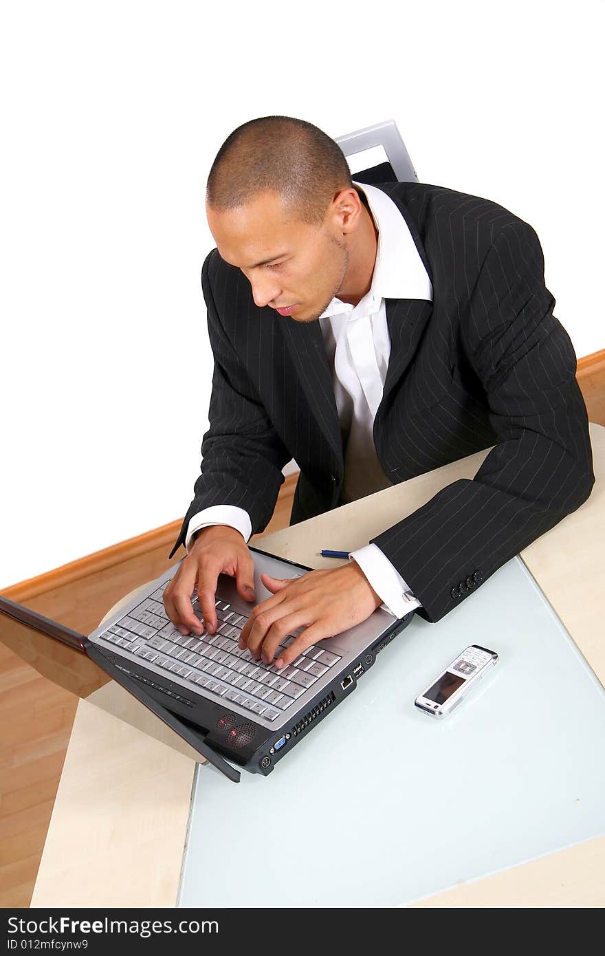 A young businessman sitting by desk at office working on the laptop with cellphone on the table.Isolated over white. A young businessman sitting by desk at office working on the laptop with cellphone on the table.Isolated over white.