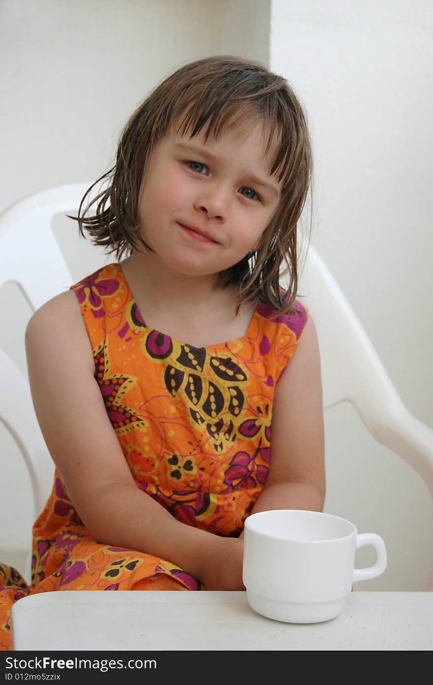 Portrait of the little girl In an orange dress sitting at a table and a white cup. Portrait of the little girl In an orange dress sitting at a table and a white cup