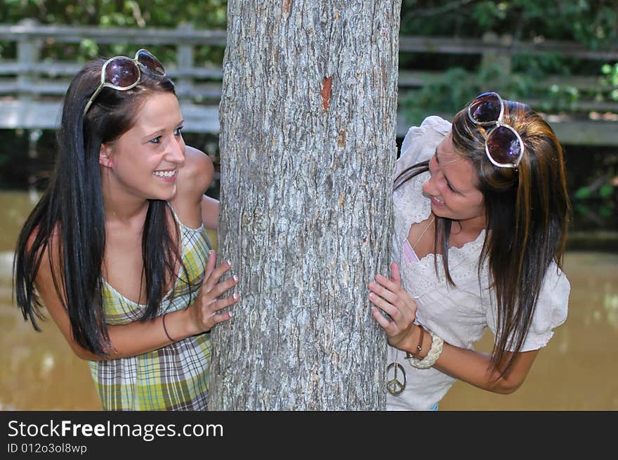 Two young ladies peeks around a tree with a lake and foot bridge in the background. Two young ladies peeks around a tree with a lake and foot bridge in the background.