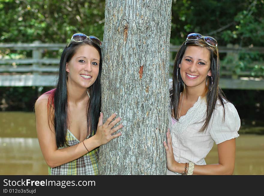 Two young ladies peeks around a tree with a lake and foot bridge in the background. Two young ladies peeks around a tree with a lake and foot bridge in the background.