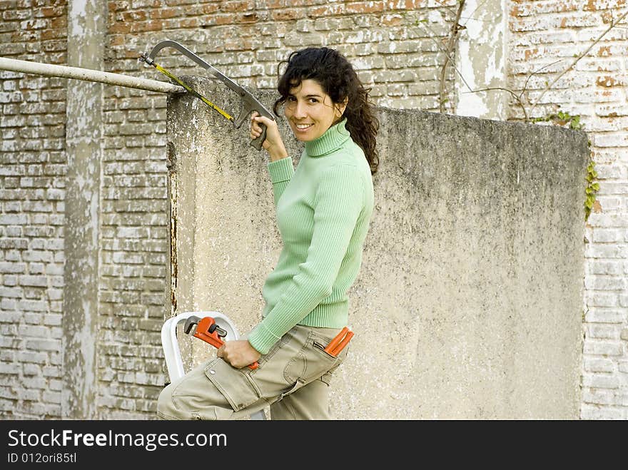 Smiling woman standing on a stool holding a wrench and a saw. Horizontally framed photo. Smiling woman standing on a stool holding a wrench and a saw. Horizontally framed photo.