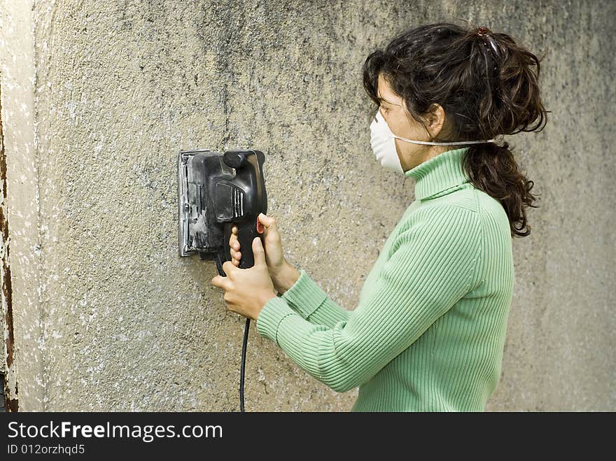 Woman wearing a paint mask uses an electric sander to sand a wall. Horizontally framed photo. Woman wearing a paint mask uses an electric sander to sand a wall. Horizontally framed photo.