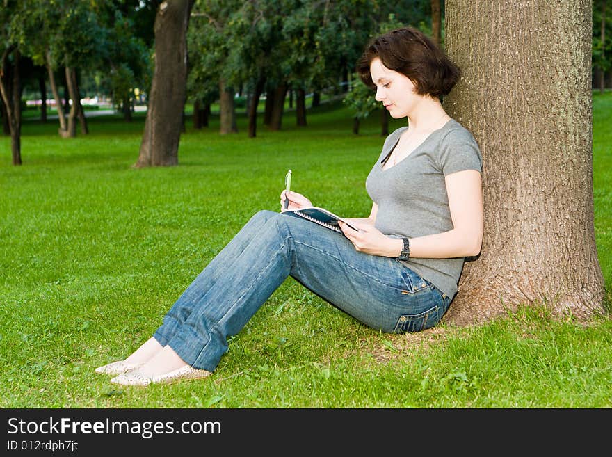 Girl sits under a tree in park and writes to notebook. Girl sits under a tree in park and writes to notebook