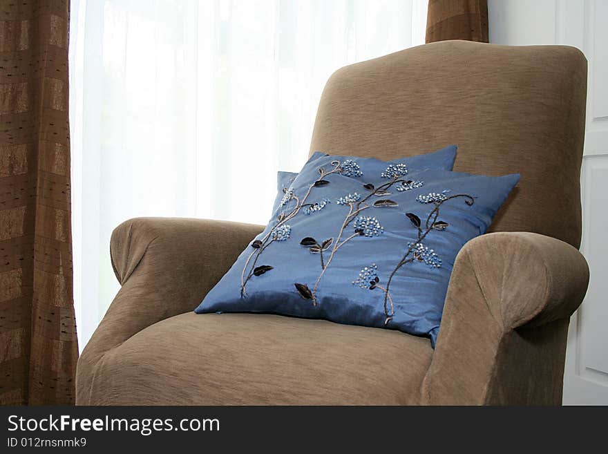 A brown chair with blue pillows sits by a window. A brown chair with blue pillows sits by a window.