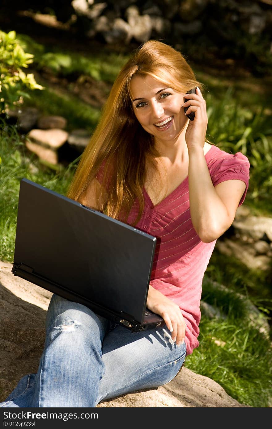 Happy young student using laptop and speaking by phone in natural environment.