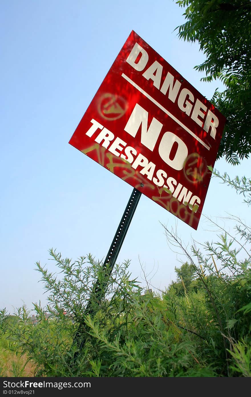 A shaded, red, metal sign reading Danger No Trespassing with graffiti markings on it, leaning out of the ground with green weeds, plants, and tree leaves behind it, against a clear blue sky