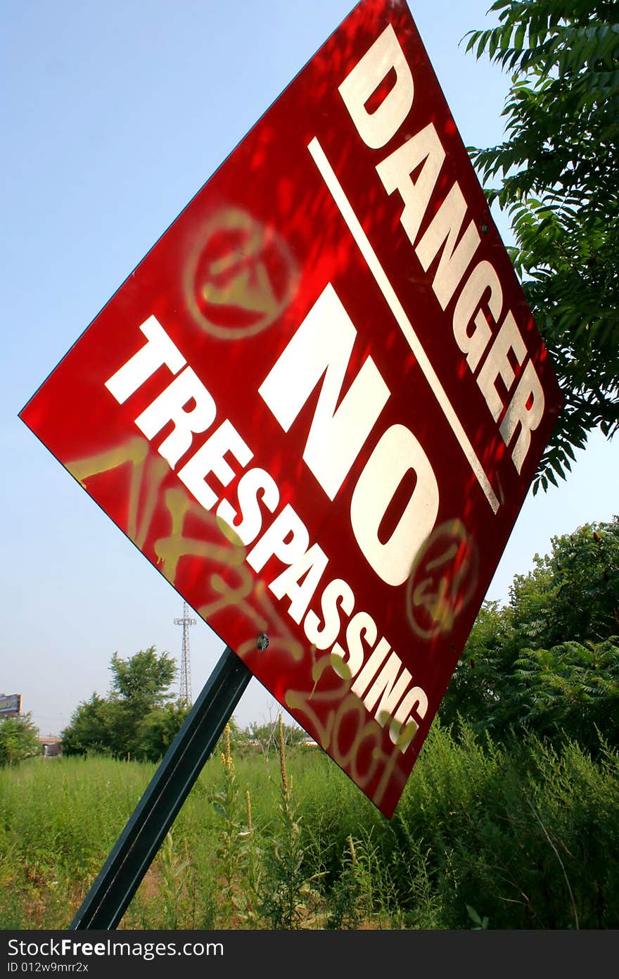 A shaded, red, metal sign reading Danger No Trespassing with graffiti markings on it, leaning out of the ground with a field of green weeds, plants, and tree leaves behind it, against a clear blue sky