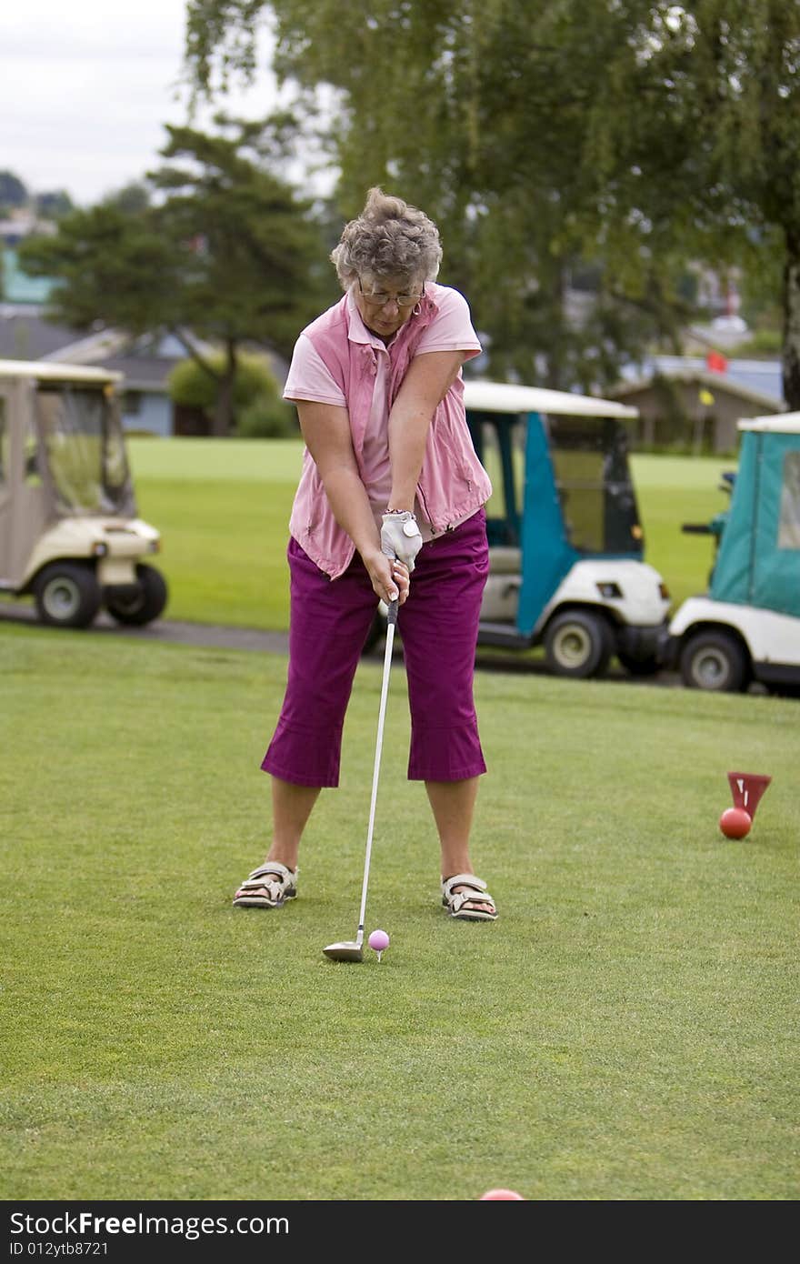 Elderly woman teeing off with golf carts in the background. Vertically framed photo. Elderly woman teeing off with golf carts in the background. Vertically framed photo.