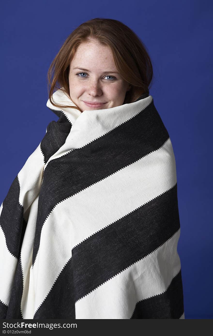 Young beautiful red headed woman with freckels and blue eyes wraps herself in a striped blanket.  She is against a deep blue background. Young beautiful red headed woman with freckels and blue eyes wraps herself in a striped blanket.  She is against a deep blue background.