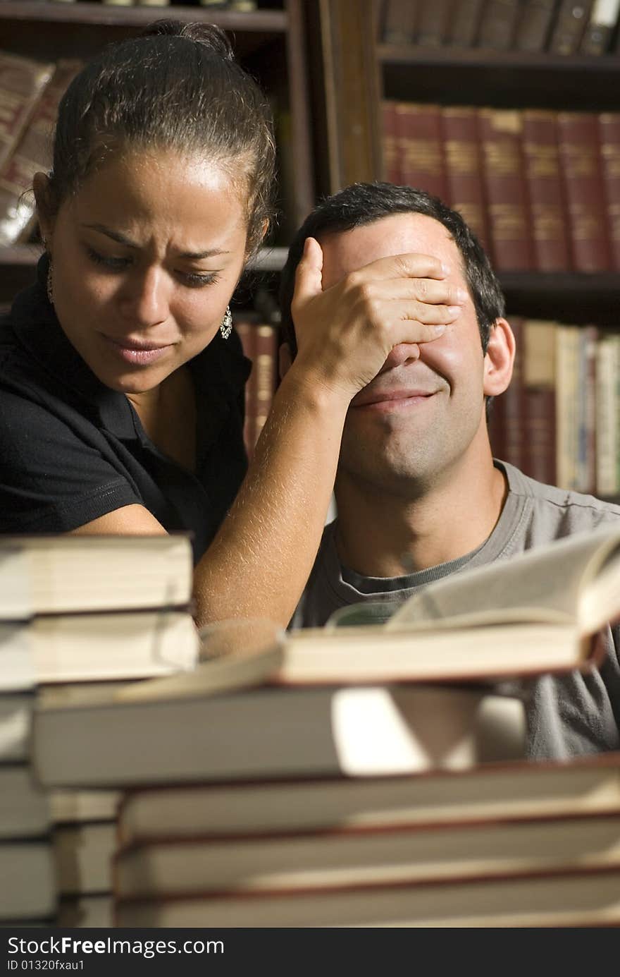 Woman covers man's eyes while studying in library. Vertically framed photo. Woman covers man's eyes while studying in library. Vertically framed photo.