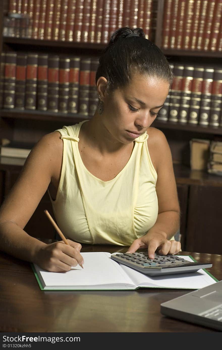 Woman sits at a table in the library. She is studying and has a calculator resting on her book.  Vertically framed photo. Woman sits at a table in the library. She is studying and has a calculator resting on her book.  Vertically framed photo.