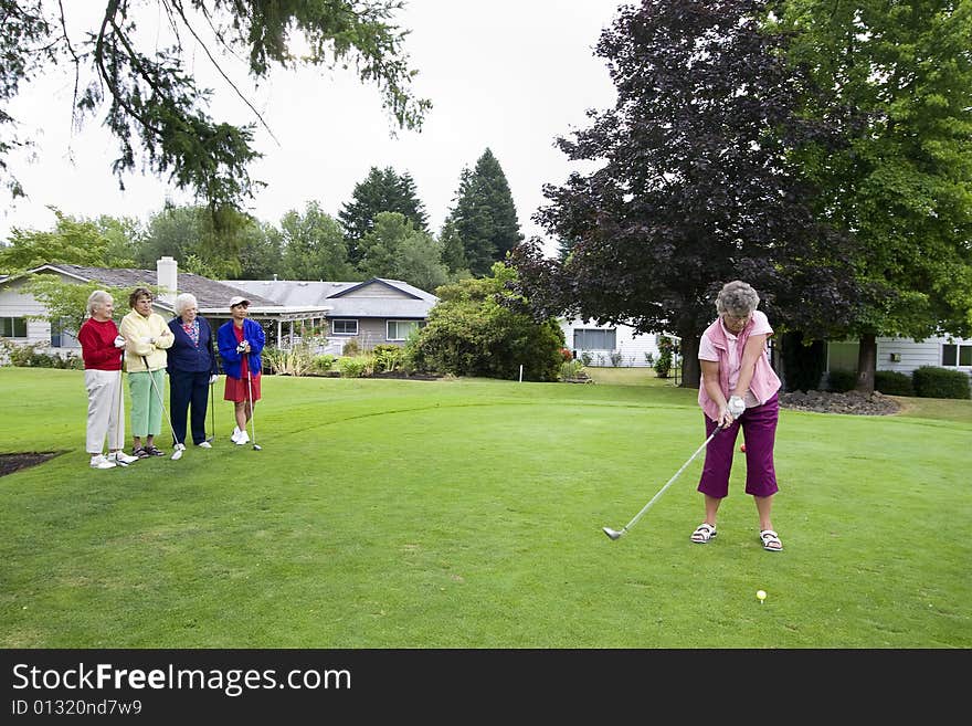 Elderly woman teeing off on the golf course as her four friends watch. Horizontally framed photo.